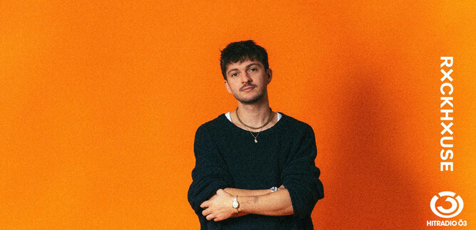 A young man is standing in front of an orange background. He is wearing a black top and has a relaxed pose. | © Mario Wallner