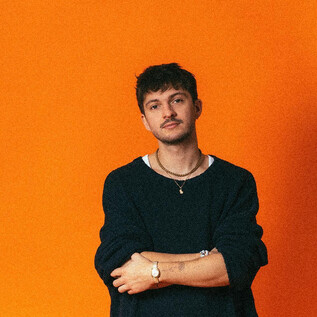 Ein junger Mann steht vor einem orangefarbenen Hintergrund. Er trägt ein schwarzes Oberteil und hat eine entspannte Pose. | © Mario Wallner