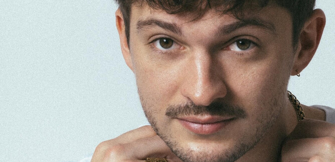 Ein junger Mann mit kurzen lockigen Haaren und einem leichten Bart. Er trägt ein weißes T-Shirt und schmückt sich mit mehreren Schmuckstücken. | © Mario Wallner