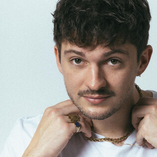 A young man with short curly hair and a light beard. He is wearing a white T-shirt and adorns himself with several pieces of jewelry. | © Mario Wallner