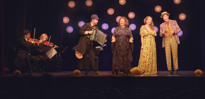 A colorful stage performance featuring five artists. Two female singers and three musicians, including an accordion player, are in the spotlight with bright background lights. | © Stefan Liewehr