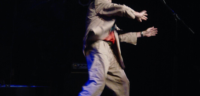 Ein Künstler auf der Bühne führt eine Performance auf. Er trägt einen Anzug und einen Hut, während bunte Lichter leuchten. | © Roswitha Pross