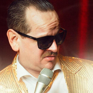 Ein Mann in einer goldenen Jacke mit Sonnenbrille hält ein Mikrofon. Im Hintergrund leuchtet eine glamouröse Kulisse. | © Thomas Steineder