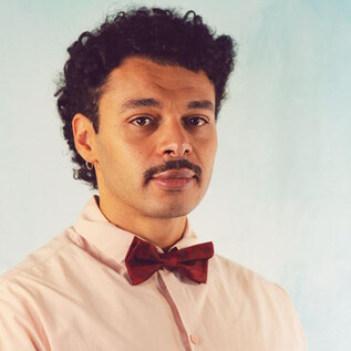 Ein Mann mit lockigem Haar trägt ein rosa Hemd und eine bordeauxfarbene Fliege. Der Hintergrund ist hell und einfarbig. | © Isabella Aust