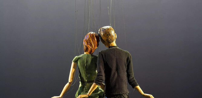 Eine Figur oder Marionette eines Paares sitzt gemeinsam, von Fäden gehalten. Der Hintergrund ist dunkel und unauffällig. | © Bernhard Müller