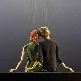 Postava nebo loutka páru sedí společně, držena nitěmi. Pozadí je tmavé a nenápadné. | © Bernhard Müller