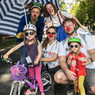 Veselá skupina lidí s červenými nosy a slunečními brýlemi, kteří pózují venku. Děti na kolech a dospělí s barevnými doplňky si užívají den. | © Rote Nasen