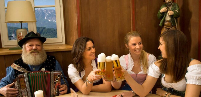A traditional Bavarian scene with a man in traditional attire playing an accordion, and three women in dirndls. They are clinking beer glasses and having fun in a cozy setting. | © Stiegl-Brauwelt