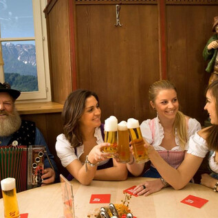 A traditional Bavarian scene with a man in traditional attire playing an accordion, and three women in dirndls. They are clinking beer glasses and having fun in a cozy setting. | © Stiegl-Brauwelt