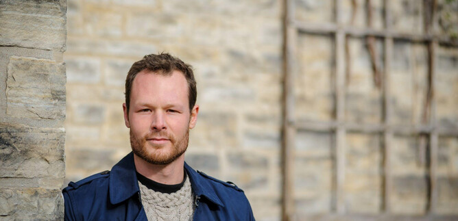 Un uomo con i capelli biondi è in piedi accanto a un muro di pietra. Indossa un mantello blu e guarda direttamente nella fotocamera. | © Bill Mccarthy