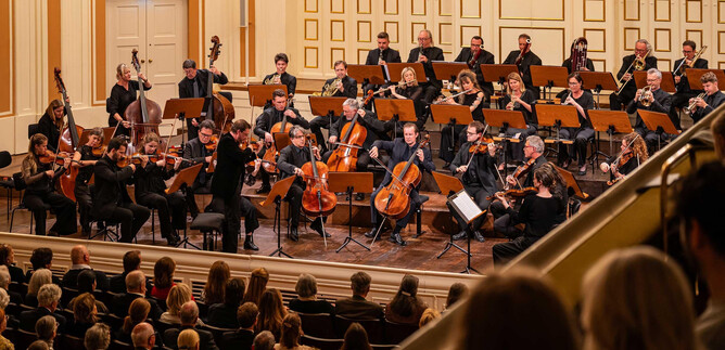 Un'orchestra suona in una sala elegante di fronte a un pubblico entusiasta. I musicisti sono riuniti sul palco e si concentrano sul direttore d'orchestra. | © Ebihara Photography