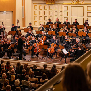 Egy zenekar játszik egy elegáns teremben egy lelkes közönség előtt. A zenészek a színpadon összegyűltek és a karmesterre összpontosítanak. | © Ebihara Photography