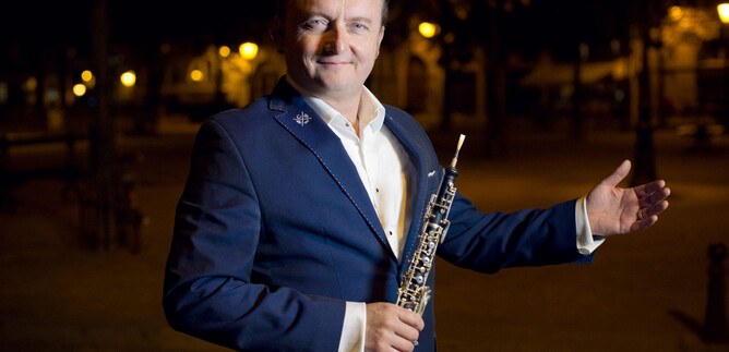 A man in a suit holds an oboe and poses smiling in a brightly lit environment. The scene radiates an artistic atmosphere. | © Jean-Baptiste-Millot
