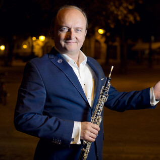 A man in a suit holds an oboe and poses smiling in a brightly lit environment. The scene radiates an artistic atmosphere. | © Jean-Baptiste-Millot