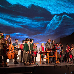 A theater performance with a group of children standing on stage and playing instruments. A projected landscape can be seen in the background. | © www.hirtenadvent.at