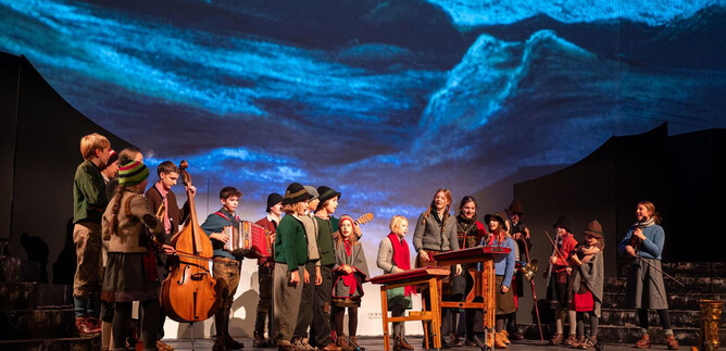 A theater performance with a group of children standing on stage and playing instruments. A projected landscape can be seen in the background. | © www.hirtenadvent.at