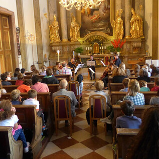 A concert hall with many spectators listening to an ensemble of four musicians. The decoration is magnificent, with golden statues and a festive ambiance.