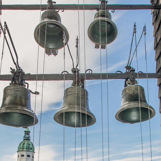 A view of bells hanging from a frame. In the background, there is a church tower with a green dome and a clear sky. | © SalzburgerLand Tourismus / Eva trifft