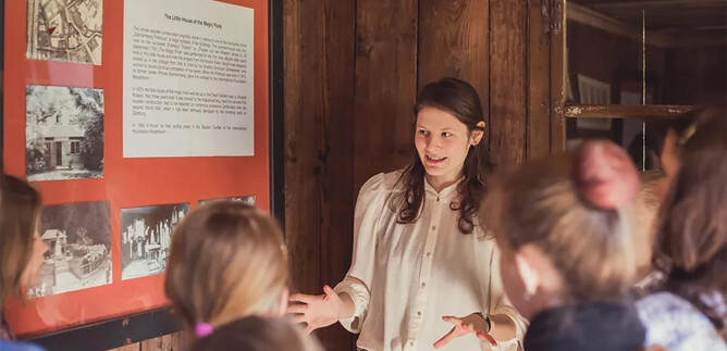 A young woman is explaining something to a group of listeners in front of a whiteboard. Historical images and texts are visible in the background. | © Eva trifft