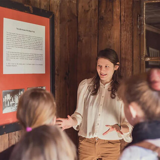 Une jeune femme explique quelque chose à un groupe d'auditeurs devant un tableau. En arrière-plan, des images et des textes historiques sont visibles. | © Eva trifft