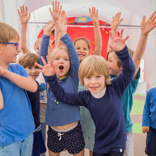 A group of happy children stands together and raises their hands in the air. They are wearing casual clothes in various shades of blue and are laughing.