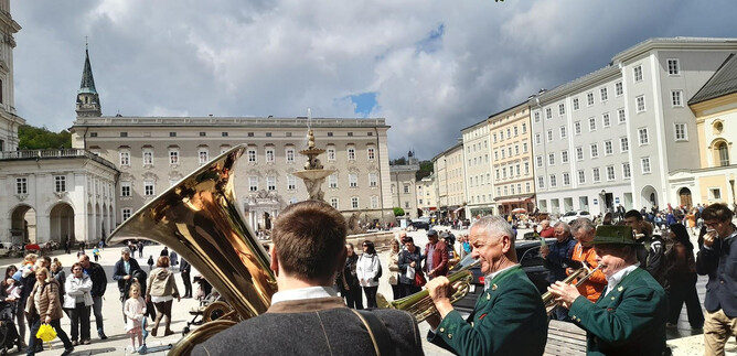 A group of musicians is playing in a square, surrounded by numerous spectators. In the background, historical buildings and a partially cloudy sky can be seen. | © Salzburger ViolksLiedWerk