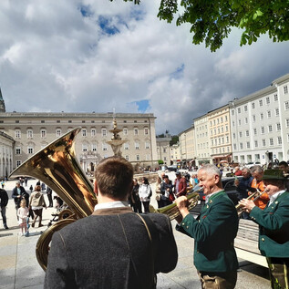 A group of musicians is playing in a square, surrounded by numerous spectators. In the background, historical buildings and a partially cloudy sky can be seen. | © Salzburger ViolksLiedWerk