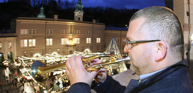 A man is playing trumpet on a balcony. In the background, festively illuminated stalls and crowds of people can be seen. | © www.christkindlmarkt.co.at, Salzburg