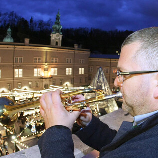 Ein Mann spielt Trompete auf einem Balkon. Im Hintergrund sind festlich beleuchtete Stände und Menschenmengen zu sehen. | © www.christkindlmarkt.co.at, Salzburg