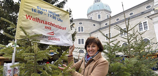 A woman stands in front of a Christmas market with a sign for a tour. In the background, there are fir trees and a historic building visible. | © salzburg-experience.at