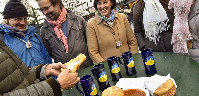 Eine fröhliche Gruppe von Menschen steht um einen Tisch mit Essen und Getränken. Auf dem Tisch liegen Brötchen und ein Teller mit einer warmen Suppe. | © salzburg-experience.at