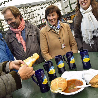 Un grupo alegre de personas está alrededor de una mesa con comida y bebidas. En la mesa hay bollos y un plato con una sopa caliente. | © salzburg-experience.at