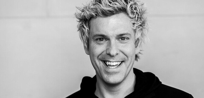 A smiling man with curly blonde hair is wearing a black hoodie and a blazer. The background is neutral and bright. | © Hoanzl Agentur GmbH