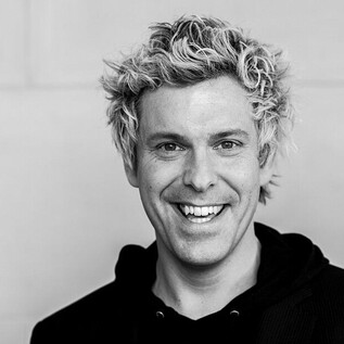 Ein lächelnder Mann mit lockigem, blondem Haar trägt einen schwarzen Hoodie und einen Blazer. Der Hintergrund ist neutral und hell. | © Hoanzl Agentur GmbH