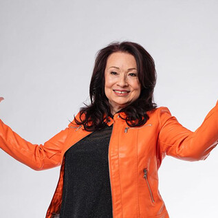 Eine lächelnde Frau in einer leuchtenden orangefarbenen Jacke hebt ihre Arme in einer einladenden Pose. Der Hintergrund ist schlicht und neutral. | © sassy.at