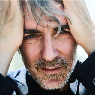 A man with gray hair is holding his head and looking directly at the camera. His facial expression appears thoughtful and intense. | © Julian Baumann