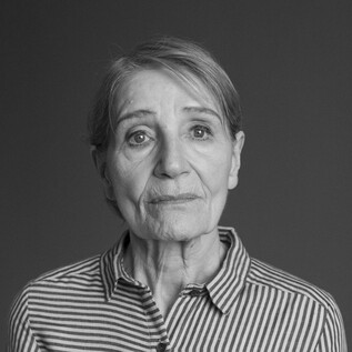 An older woman with a contemplative expression stands in front of a neutral background. She is wearing a striped shirt and has a gentle gaze. | © Armin Smailovic