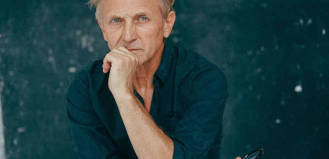 Ein nachdenklicher Mann mit blonden Haaren sitzt mit einer Hand am Kinn. Der Hintergrund ist dunkel und einfach gehalten. | © Nowy Teatr
