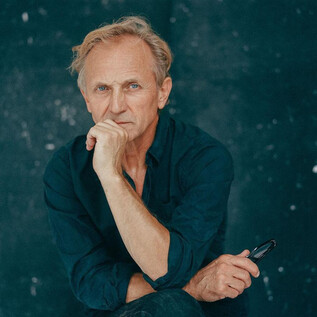 Un homme pensif aux cheveux blonds est assis avec une main sur le menton. L'arrière-plan est sombre et épuré. | © Nowy Teatr