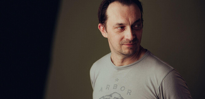 A man looks slightly to the side and wears a gray T-shirt with a pattern. The background is blurred and in neutral colors. | © Joel Heyd