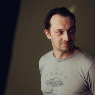 Mężczyzna lekko spogląda w bok i nosi szary T-shirt z wzorem. Tło jest nieostre i w neutralnych kolorach. | © Joel Heyd