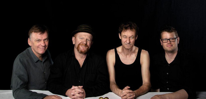 Vier Männer stehen an einem Tisch mit einer goldenen Figur in der Mitte. Sie tragen unterschiedliche Kleidung und schauen in die Kamera. | © Anna Reisinger