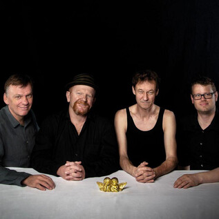 Vier Männer stehen an einem Tisch mit einer goldenen Figur in der Mitte. Sie tragen unterschiedliche Kleidung und schauen in die Kamera. | © Anna Reisinger