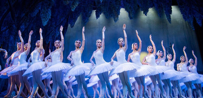 A group of dancers in white ballet costumes. They are performing a synchronized dance on a stage with a dramatic background. | © International Festival Ballet