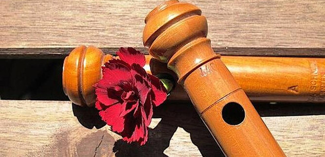 A wooden recorder lies on a wooden table. Next to it is a red carnation. | © www.salzburgervolksliedwerk.at