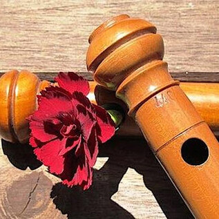 A wooden recorder lies on a wooden table. Next to it is a red carnation. | © www.salzburgervolksliedwerk.at