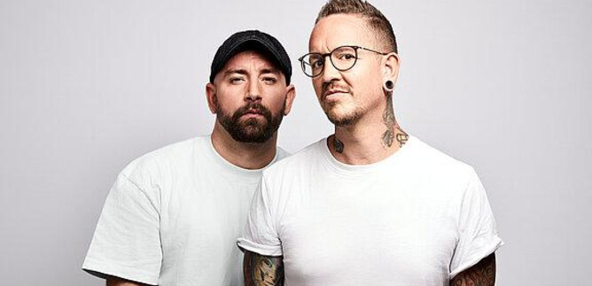 Two men are standing next to each other in front of a bright background. Both are wearing casual clothing and have tattoos. | © Pascal Riesinger