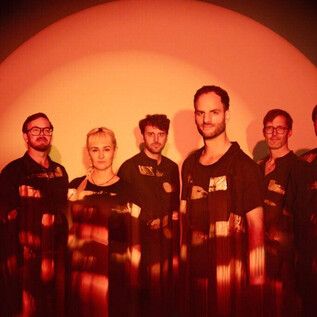 Skupina sedmi lidí stojí před oranžovým pozadím. Nosí podobné oblečení a mají různé účesy. | © Victoria Nazarova