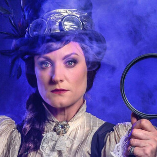 Žena v historickém oblečení drží lupu v ruce. Pozadí je mystické s modrou mlhou. | © SLT / Tobias Witzgall