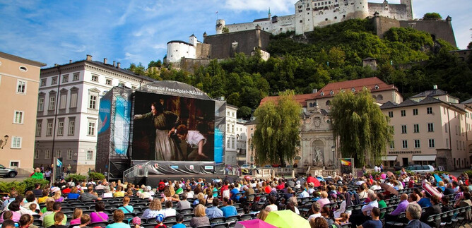 Ein großes Publikum sitzt auf Stühlen in einer Stadt, während sie eine Aufführung auf einer Leinwand ansehen. Im Hintergrund sieht man eine beeindruckende Burg auf einem Hügel. | © Siemens AG Österreich